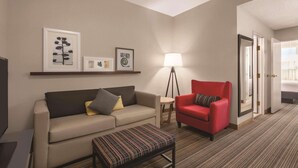 Een hotelkamer met een bank, fauteuil, klein tafeltje, lamp en een bed, met een deur naar een andere kamer.