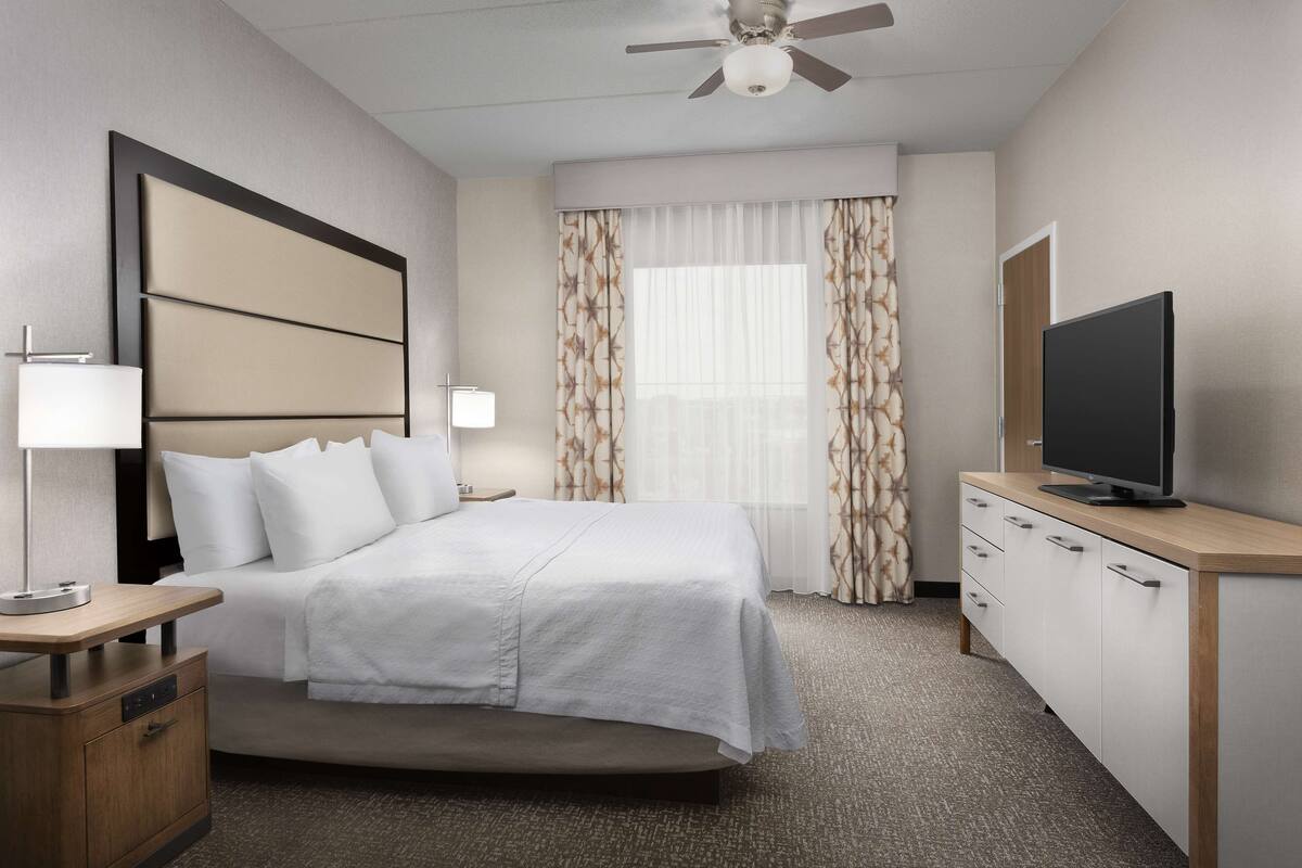 A hotel room with a bed, bedside table, lamp, TV, and a ceiling fan.