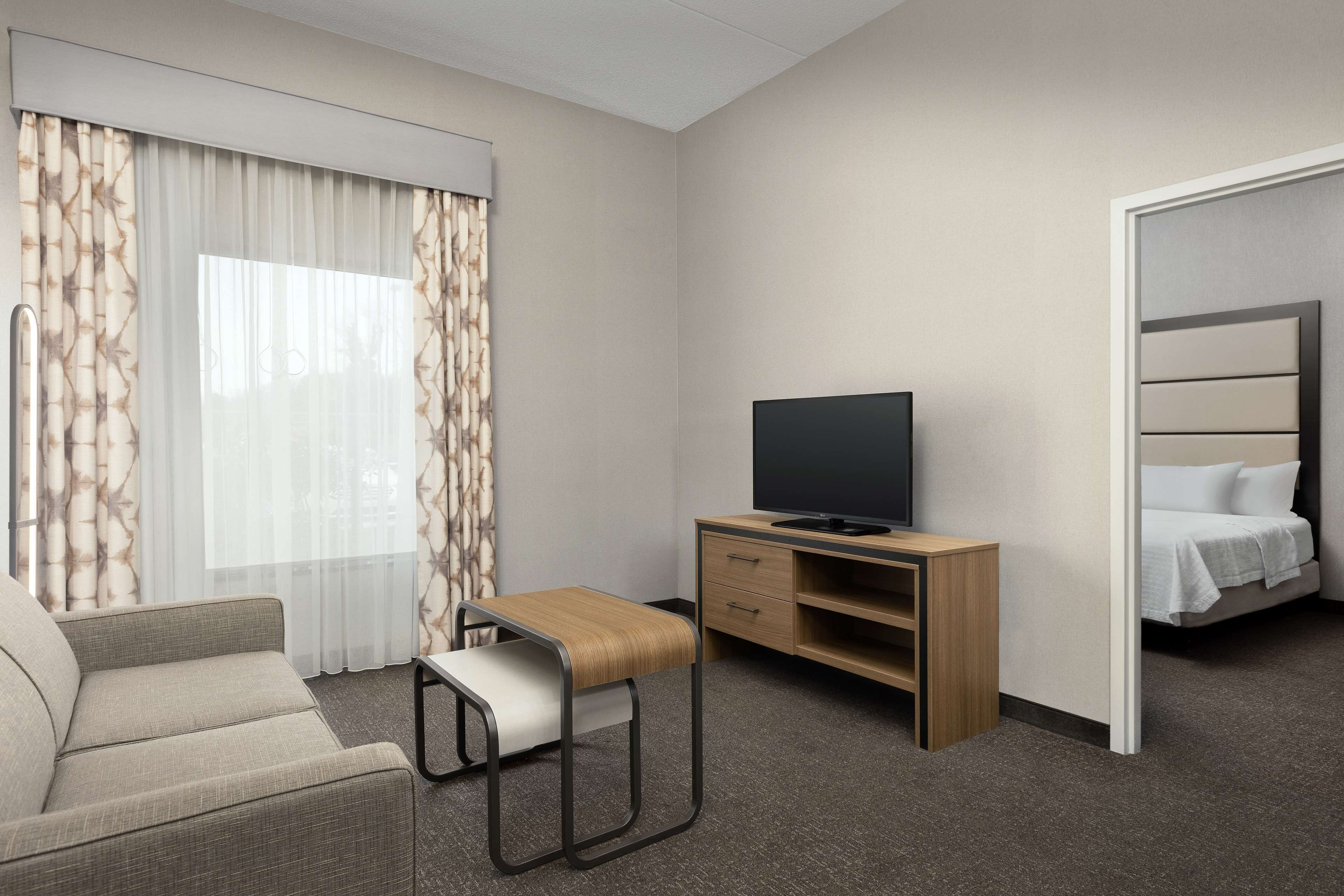 A hotel room with a sofa, a small wooden table, a TV on a wooden stand, a bed, and a window with curtains.