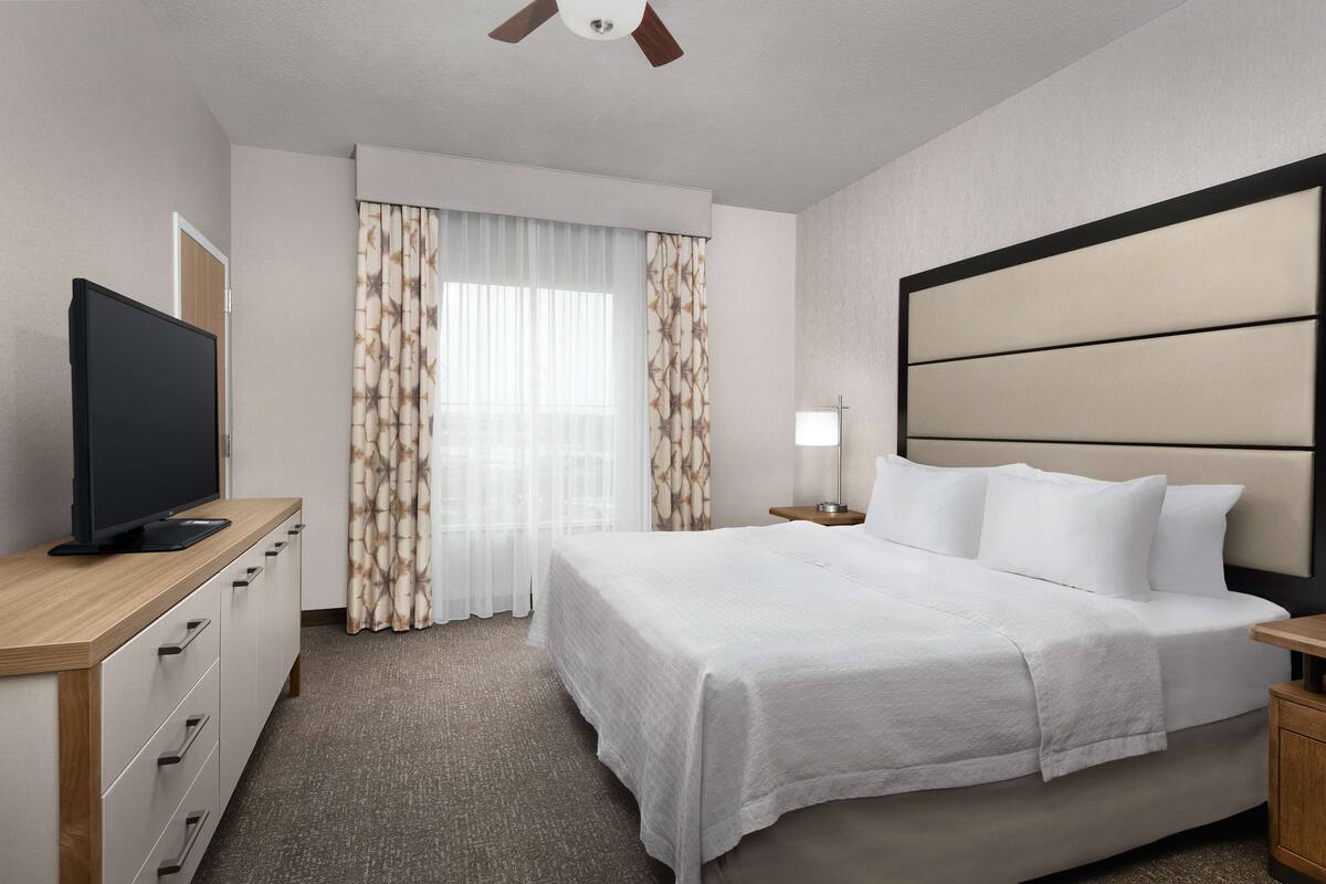 A hotel room with a large bed, a TV on a wooden dresser, a nightstand with a lamp, and a window with curtains.