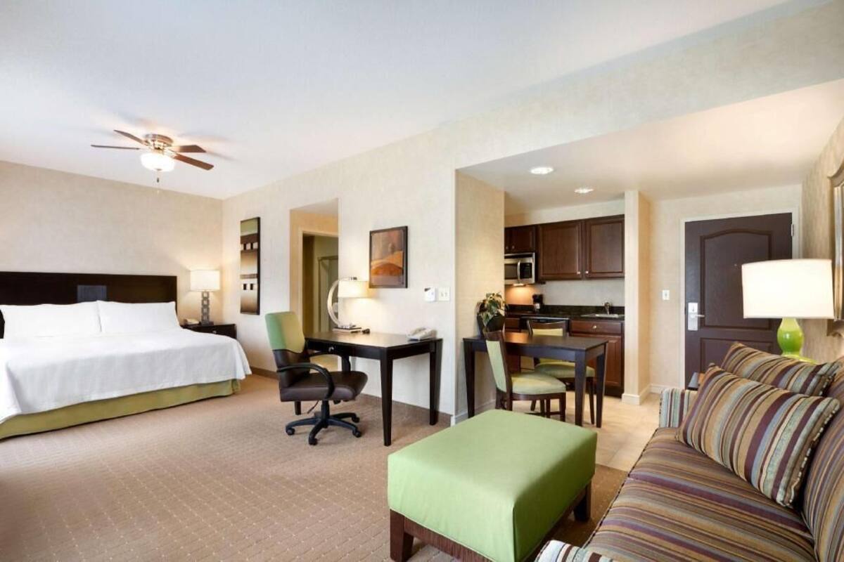 A hotel room with a bed, desk, chair, sofa, kitchenette, and a ceiling fan.