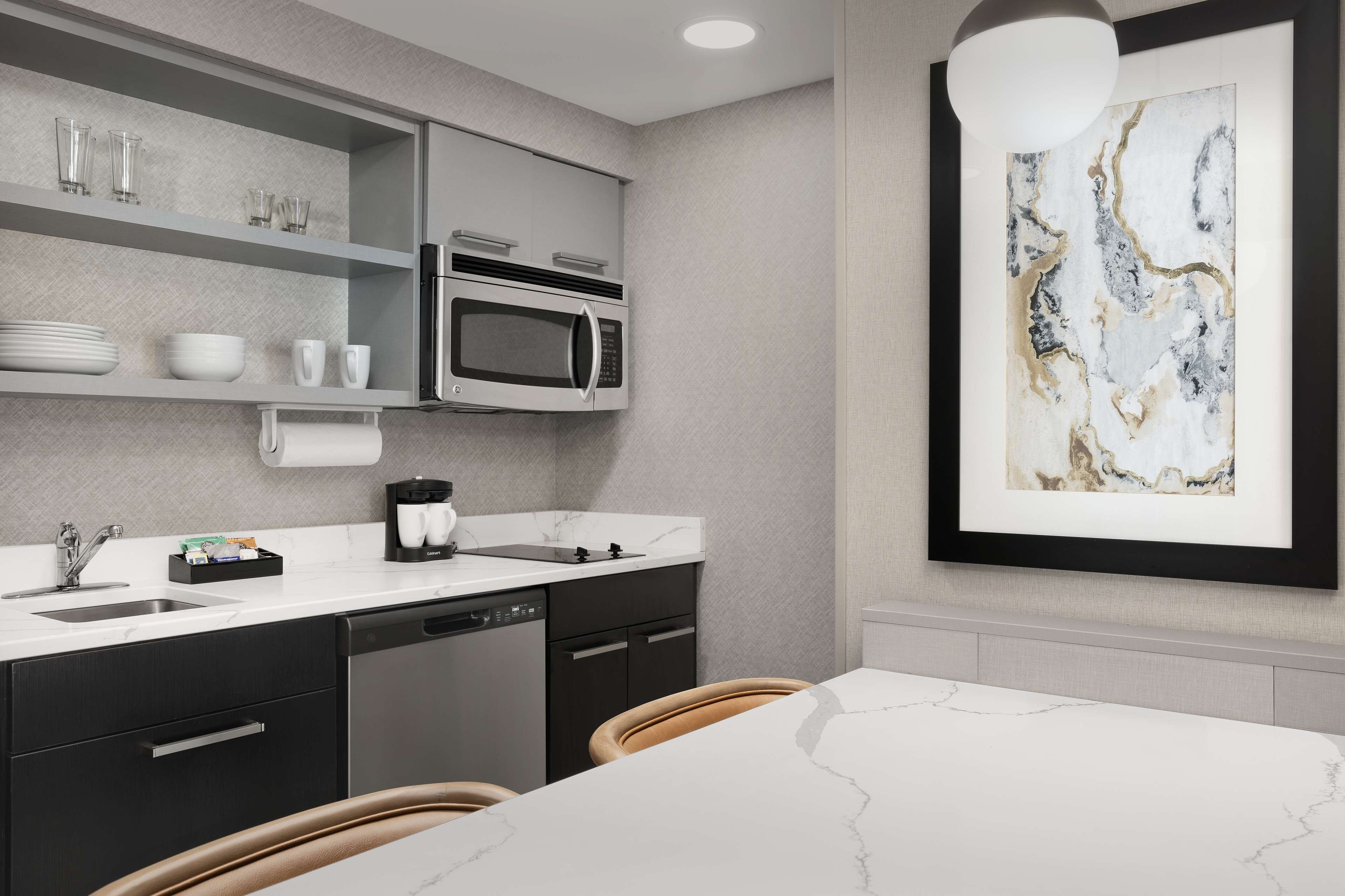 A modern kitchen with a built-in microwave, a coffee maker, and a sink. There is a dining table with chairs and a framed artwork on the wall.