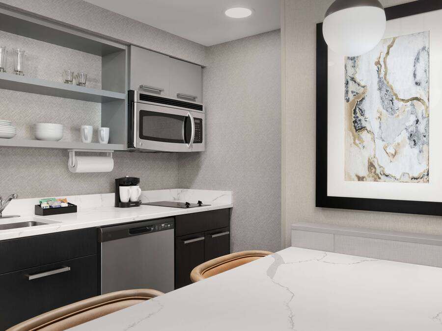 A modern kitchen with a built-in microwave, a coffee maker, and a sink. There is a dining table with chairs and a framed artwork on the wall.