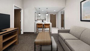 A modern hotel room with a flat-screen TV, a wooden coffee table, a sofa, a kitchen area with a dining table and chairs, and a framed picture on the wall.