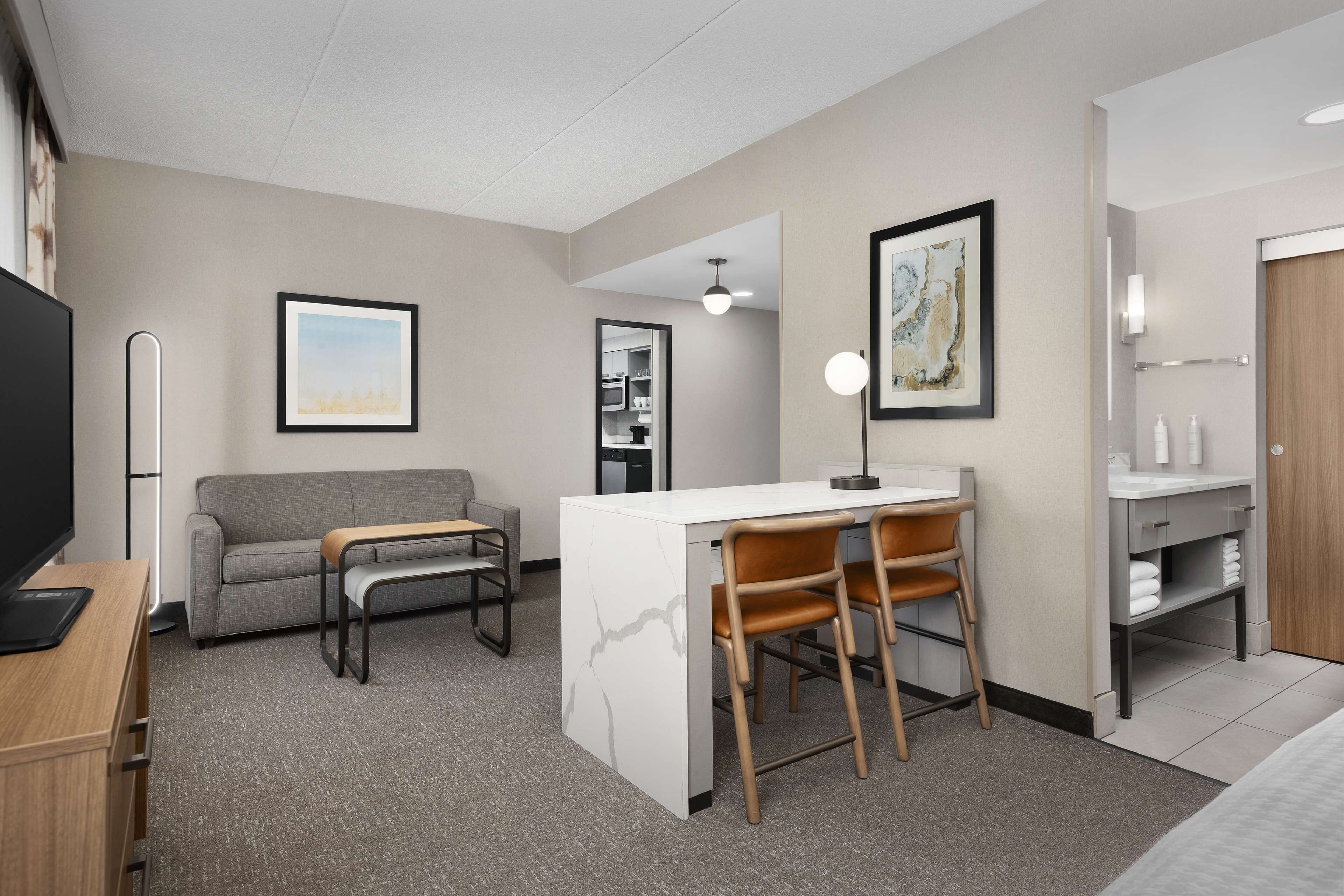 A modern hotel room with a sofa, a dining area with a table and chairs, a TV, a desk, and a bathroom with a sink and mirror.