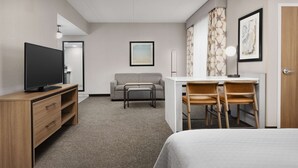 A hotel room with a bed, a TV on a wooden dresser, a desk with two chairs, a sofa, and a small table.