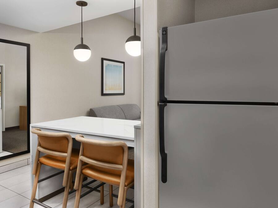 A modern kitchen with a white countertop, two pendant lights, a grey refrigerator, and wooden chairs.