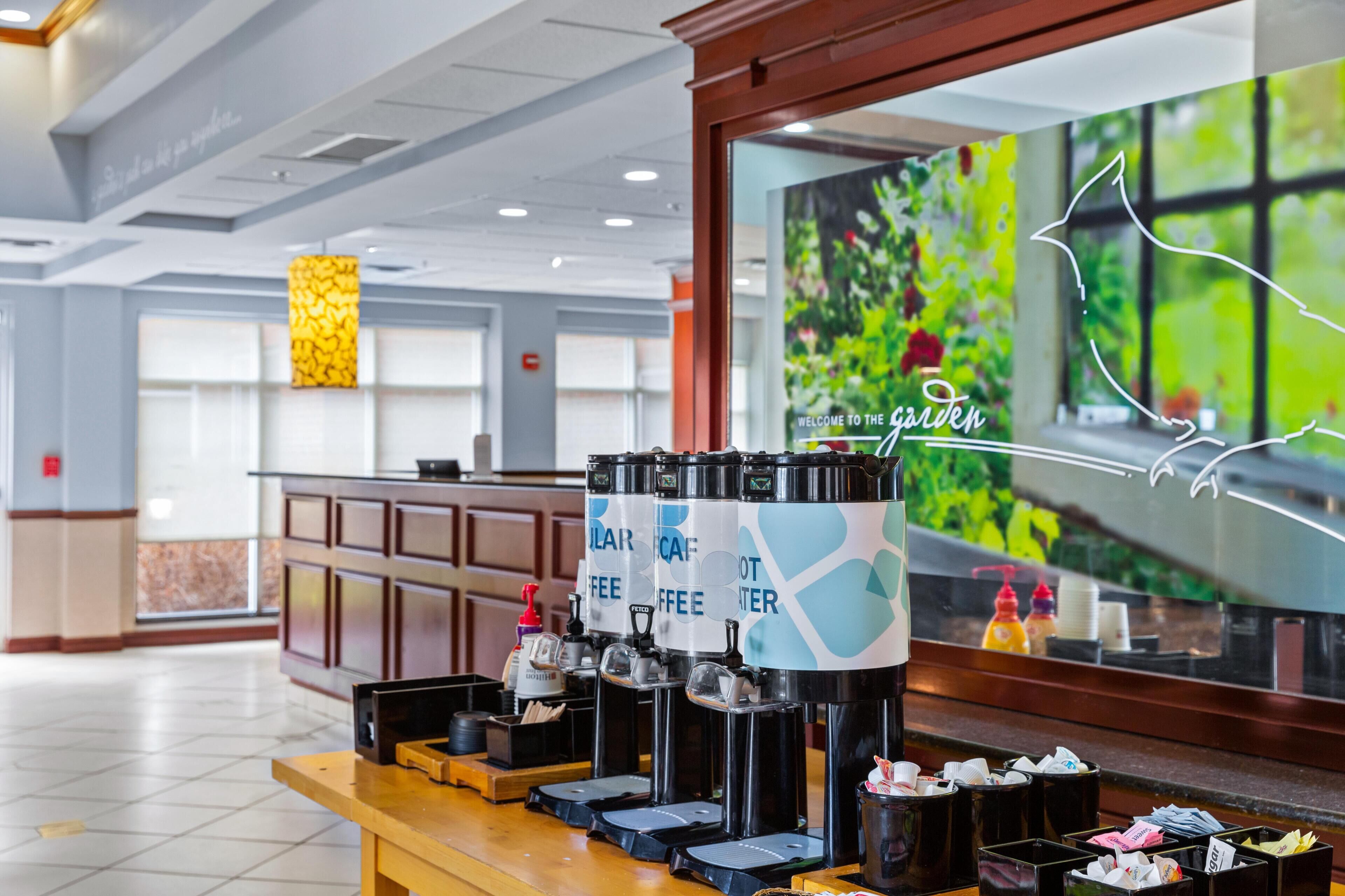 A coffee station with multiple dispensers, a reception desk, and a large window with a garden view.