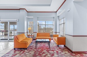 Un vestíbulo de hotel con una alfombra de diseño rojo, sofás naranjas, una mesa de centro de vidrio y ventanas amplias que ofrecen vistas al exterior.