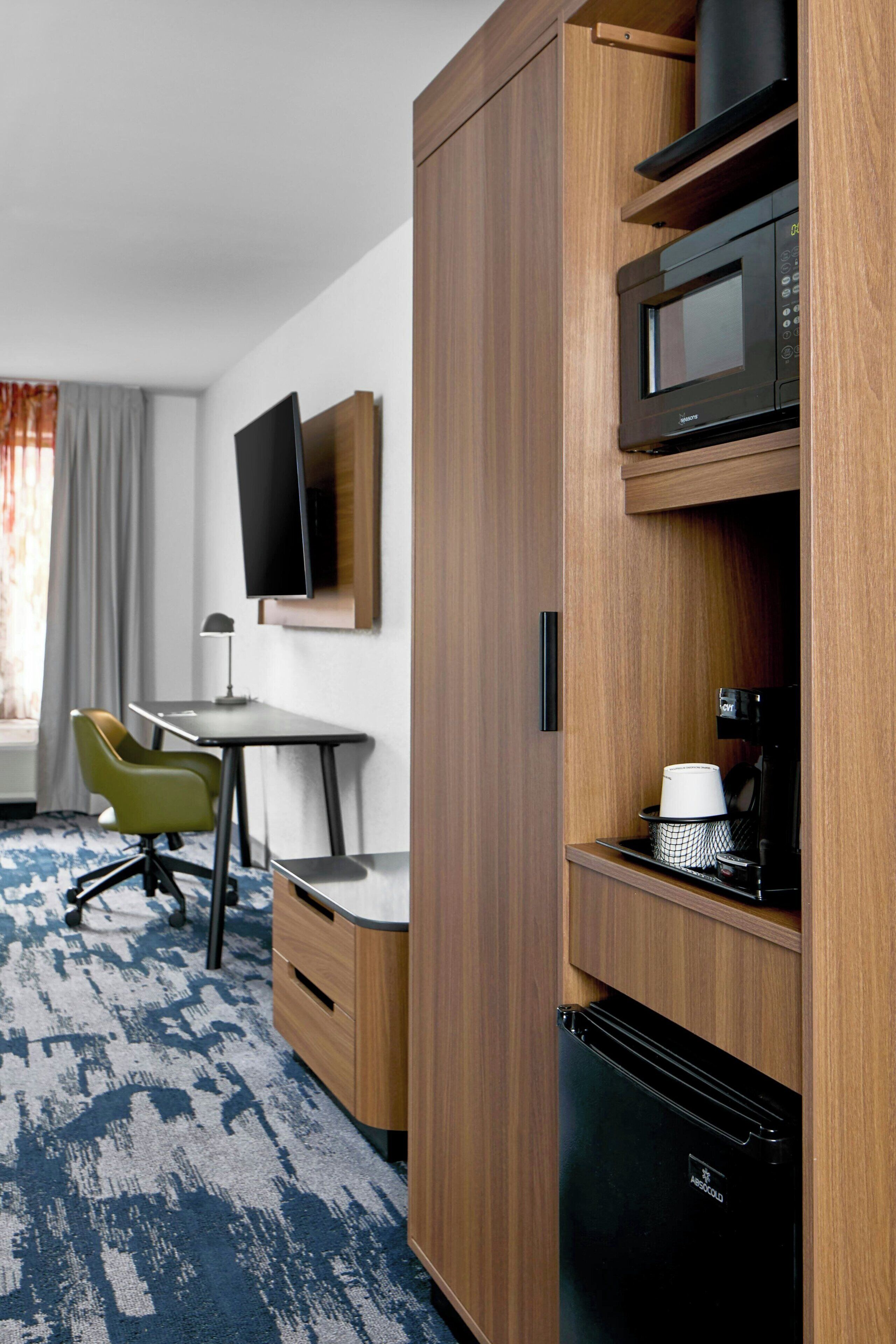 A compact hotel room with a kitchenette, a desk, a chair, a television, and a window with curtains.