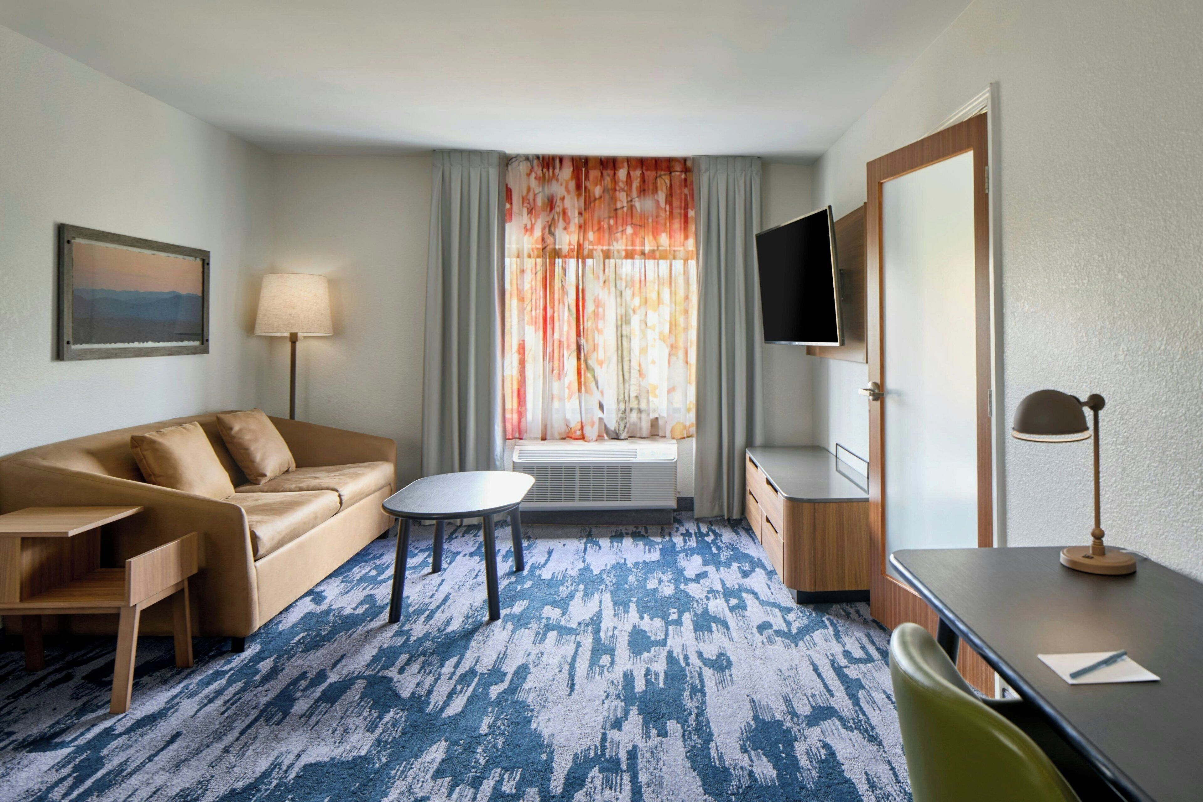 A hotel room with a sofa, a small table, a TV, a desk, and a window with curtains.