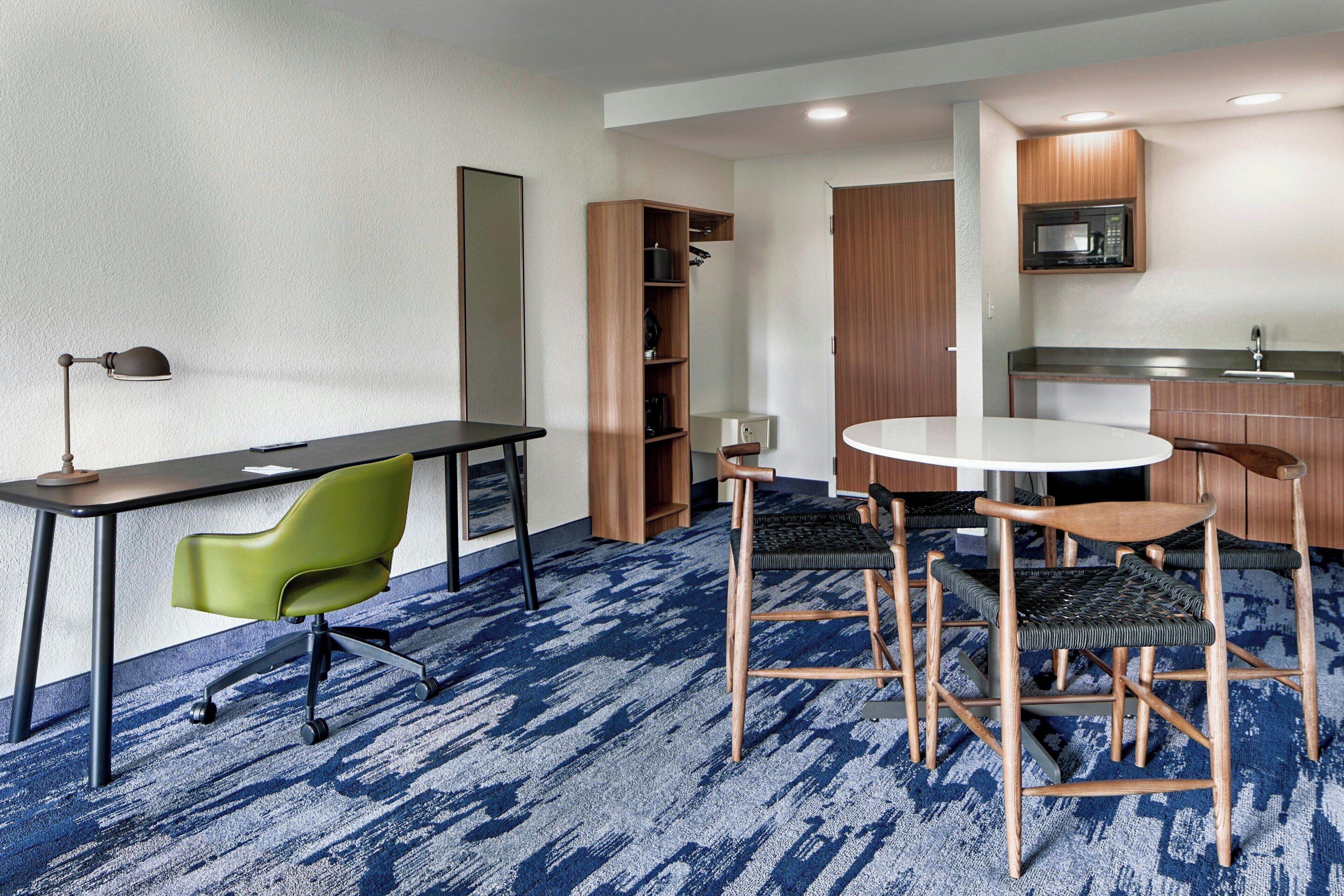 A conference room with a round table, chairs, a desk with a lamp, a bookshelf, and a kitchenette area.