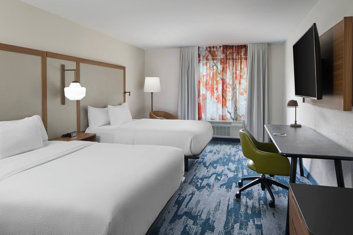 Une chambre d’hôtel avec deux lits, un bureau, une chaise, une télévision, une lampe et une fenêtre avec des rideaux.