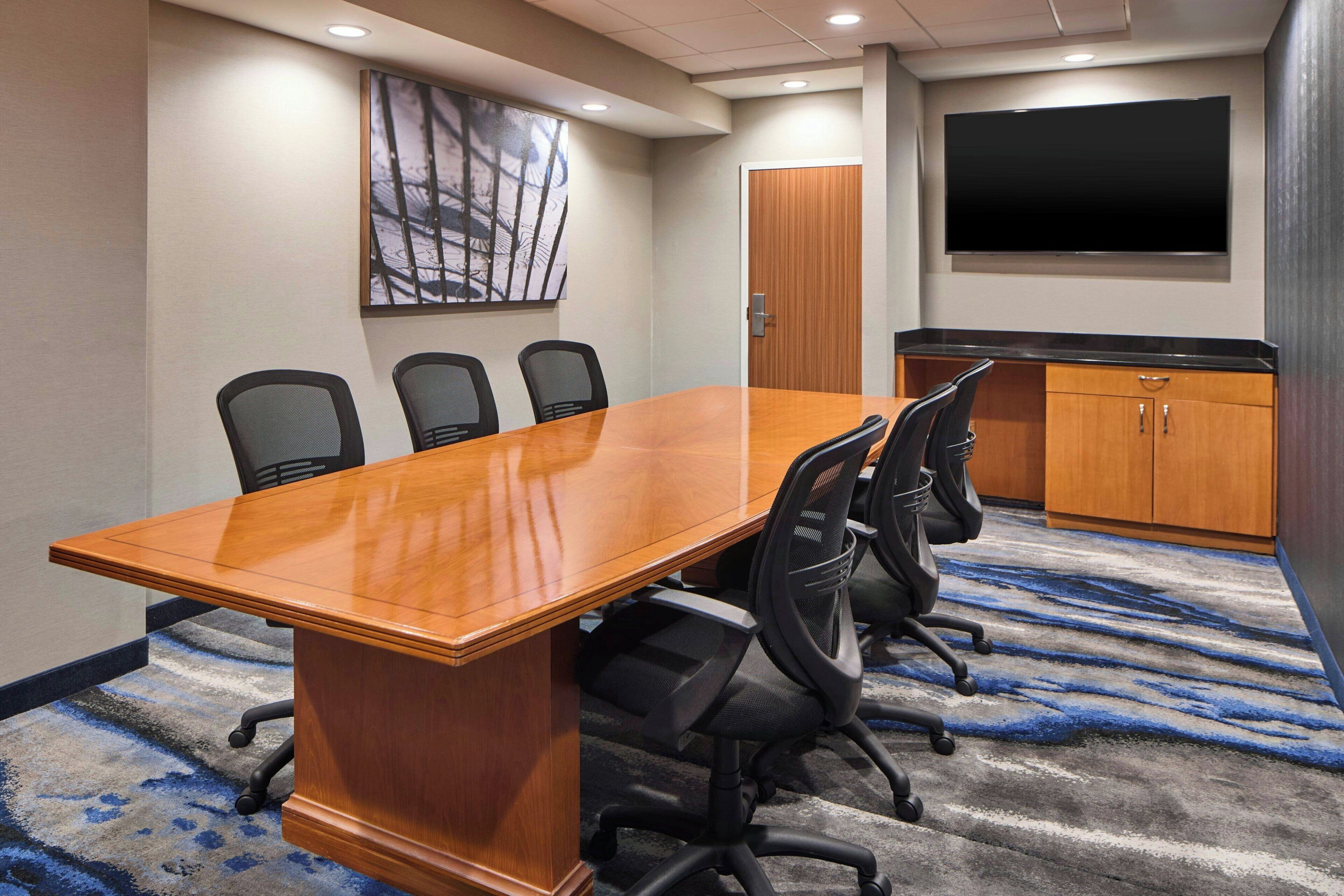 A conference room with a long wooden table, four black office chairs, a large flat-screen TV, and a wooden cabinet.
