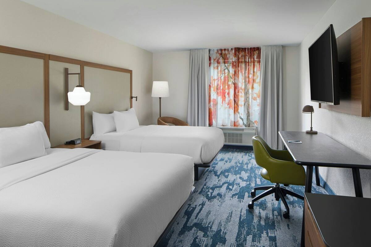 Une chambre d’hôtel avec deux lits, un bureau, une chaise, une télévision et une fenêtre avec des rideaux.