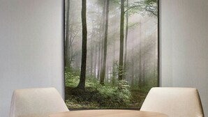 Een modern interieur met een ronde tafel, twee beige banken en een groot kunstwerk met een bosmotief aan de muur.