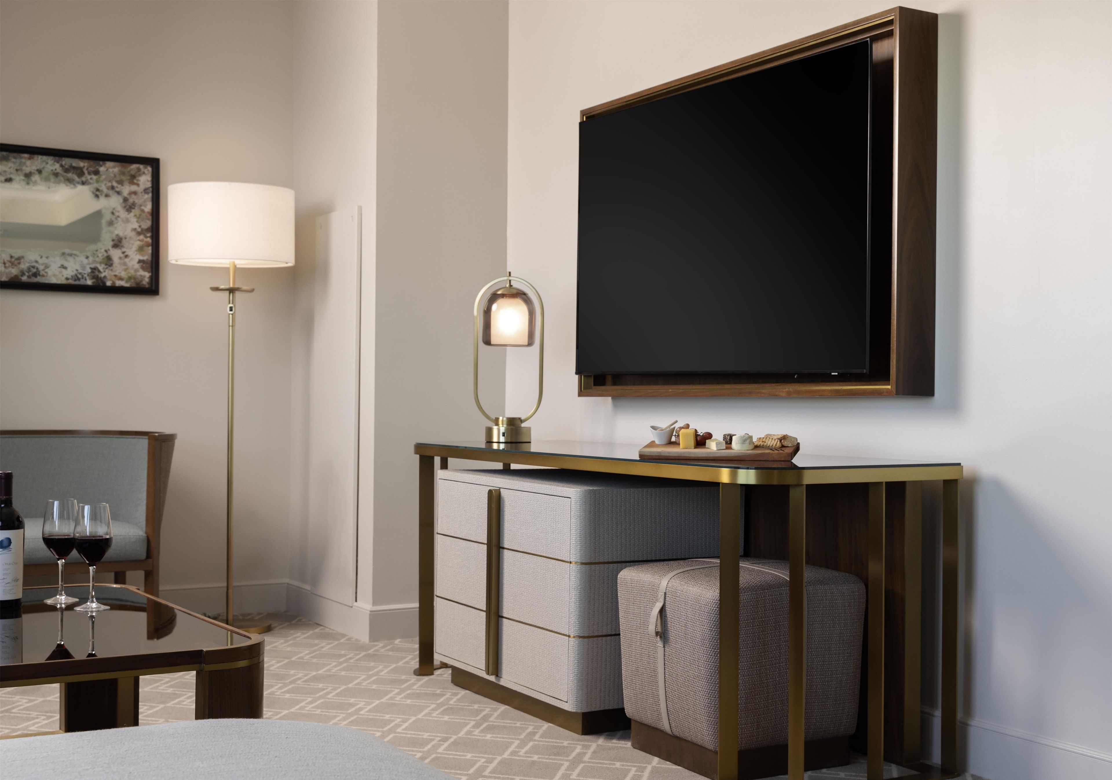 A hotel room with a flat-screen TV mounted on the wall, a small table with a lamp, a sofa, and a decorative mirror.
