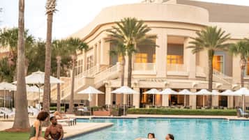 A swimming pool with palm trees and umbrellas, surrounded by a building with a curved facade and multiple balconies.