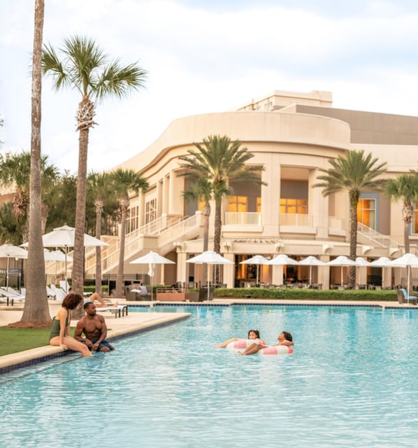 A swimming pool with palm trees and umbrellas, surrounded by a building with a curved facade and multiple balconies.