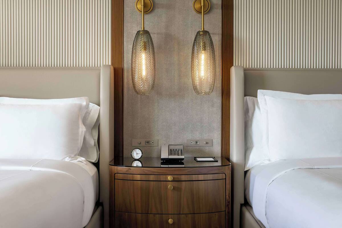 Une chambre d’hôtel avec deux lits, une table de chevet en bois sur laquelle se trouvent une horloge et un téléphone, et deux lampes suspendues.