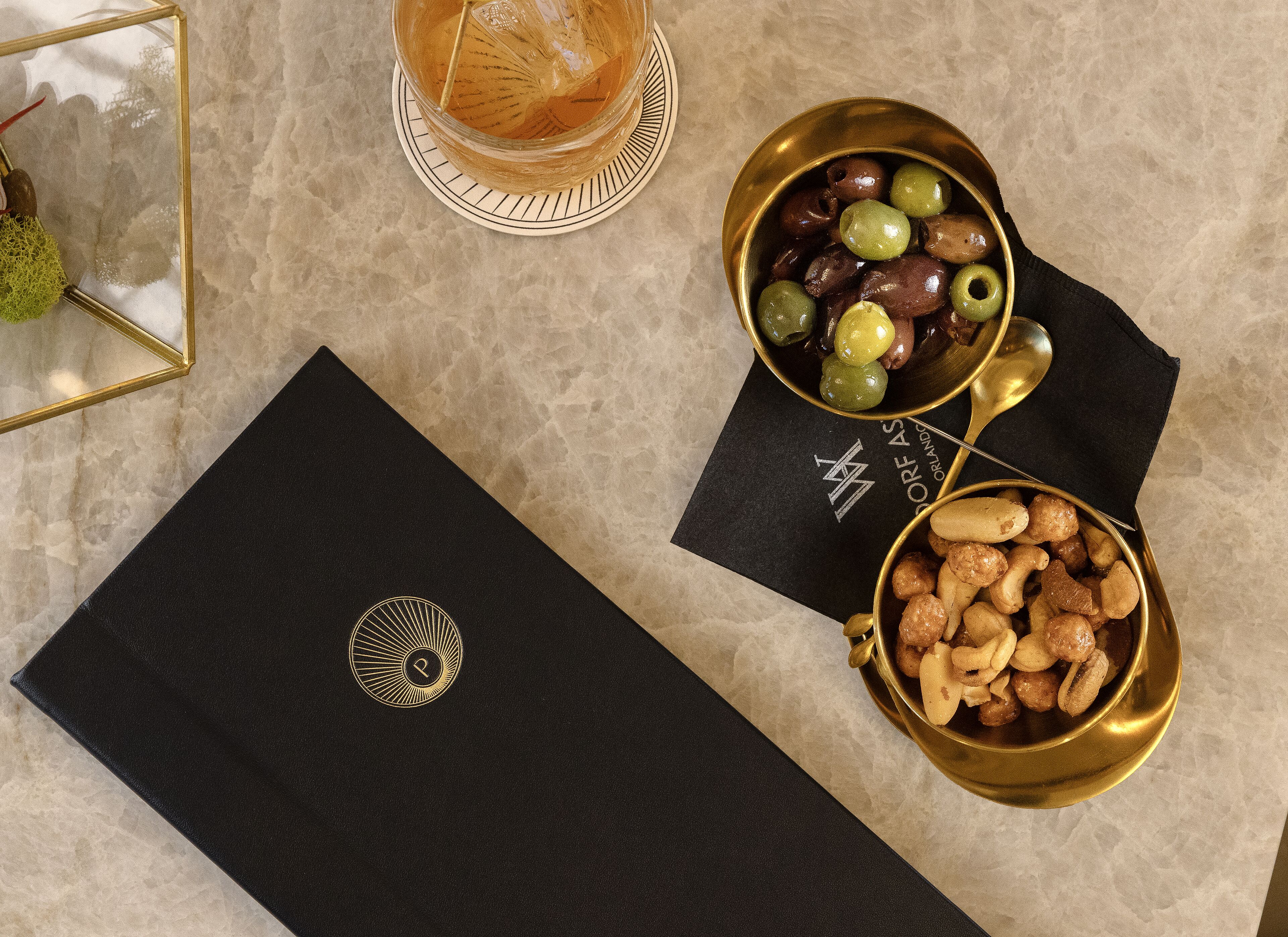 A table with a black folder, a bowl of olives, a bowl of nuts, and a glass of drink.