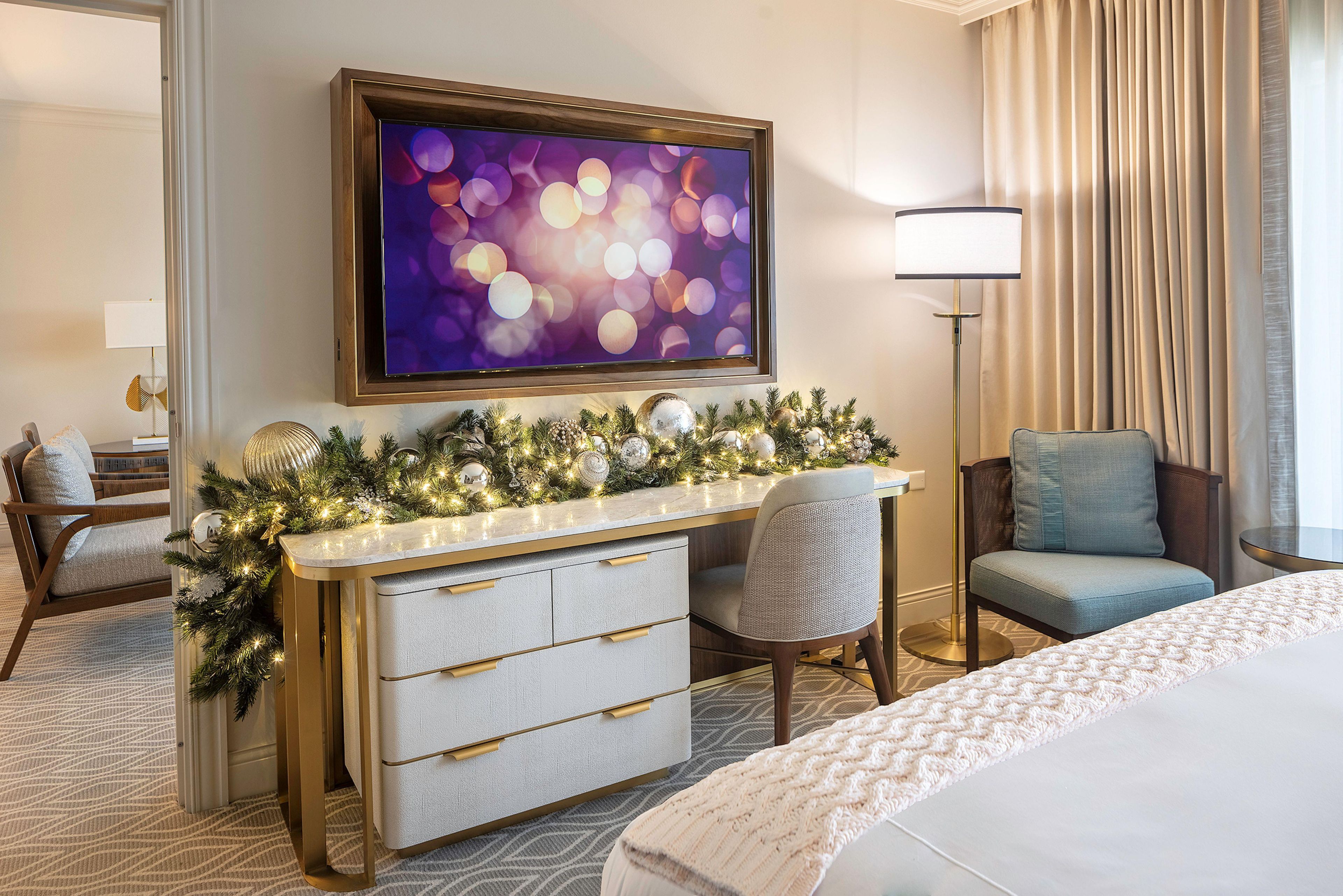 Ein Hotelzimmer mit Bett, Schreibtisch mit Schubladen, Sessel, Wandfernseher und Weihnachtsbaumdeko.