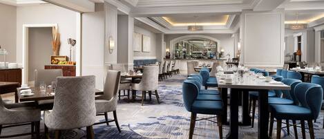 A spacious hotel dining area with multiple tables and chairs, a central chandelier, and a patterned carpet.