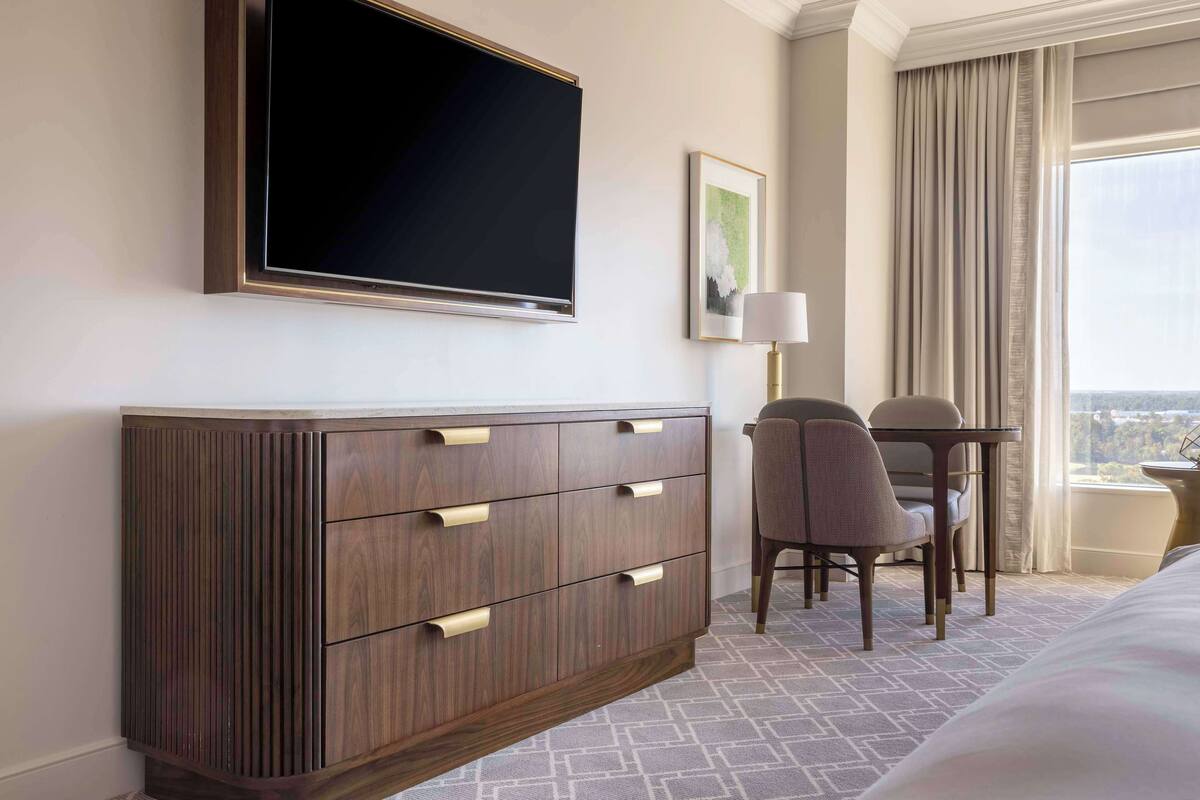 Une chambre d’hôtel dotée d’un grand téléviseur à écran plat fixé au mur, d’une commode en bois avec des tiroirs, d’un coin repas avec deux chaises et une table, et offrant une vue sur la ville par la fenêtre.