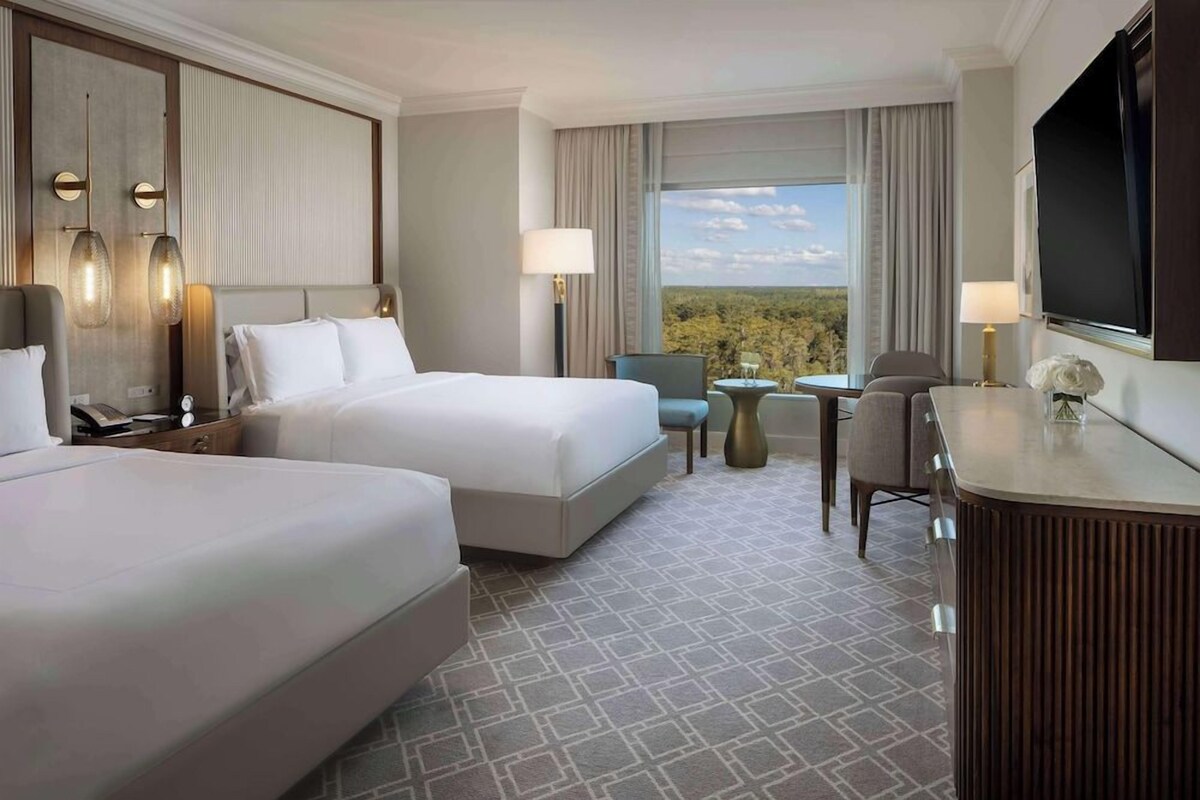 A hotel room with two beds, a desk, a chair, a TV, and a view of the outdoors.
