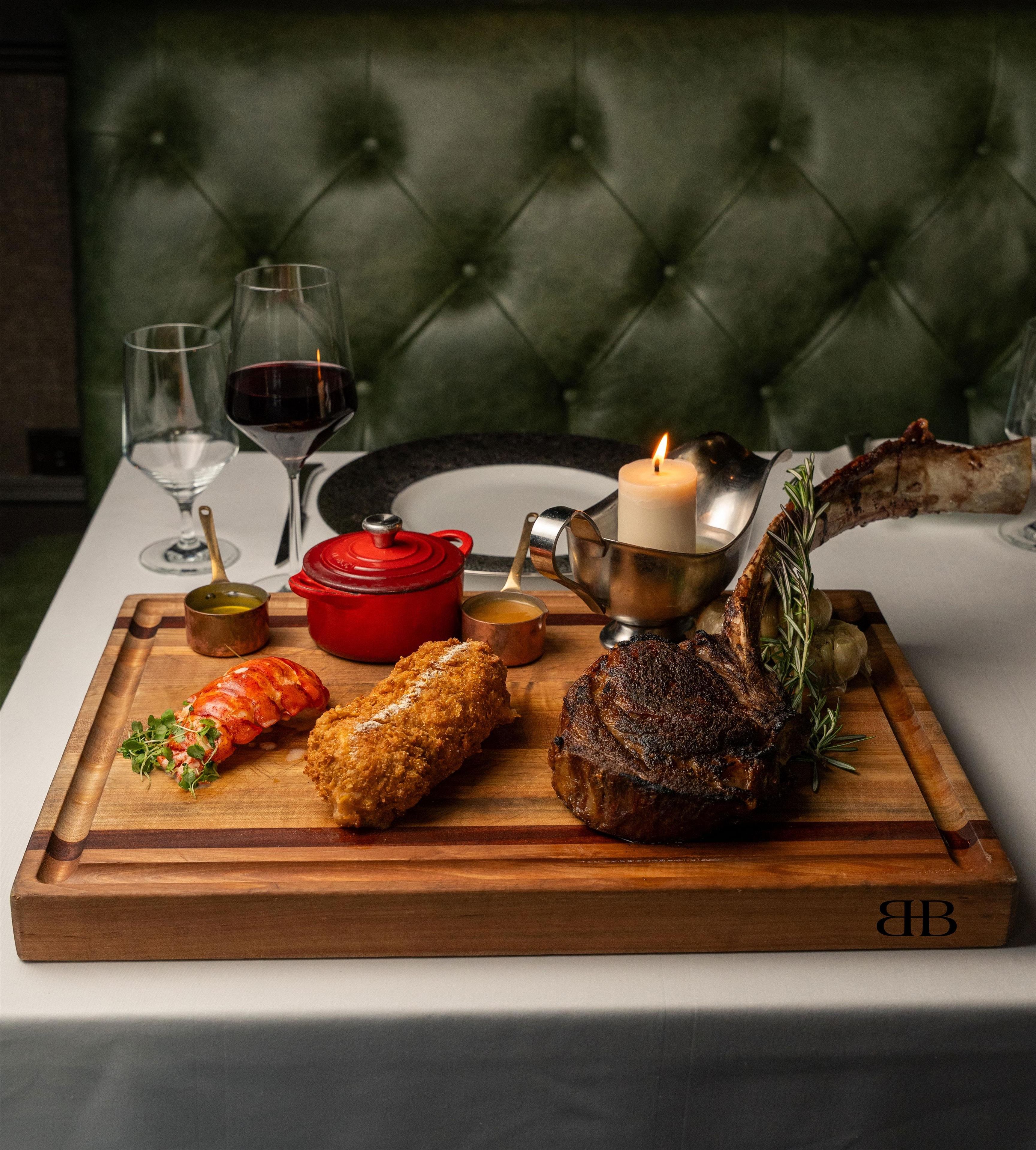 A wooden board with a lamb shank, two breaded items, a red pot, a candle, and a glass of red wine on a table.