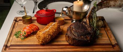 A wooden board with a lamb shank, two breaded items, a red pot, a candle, and a glass of red wine on a table.