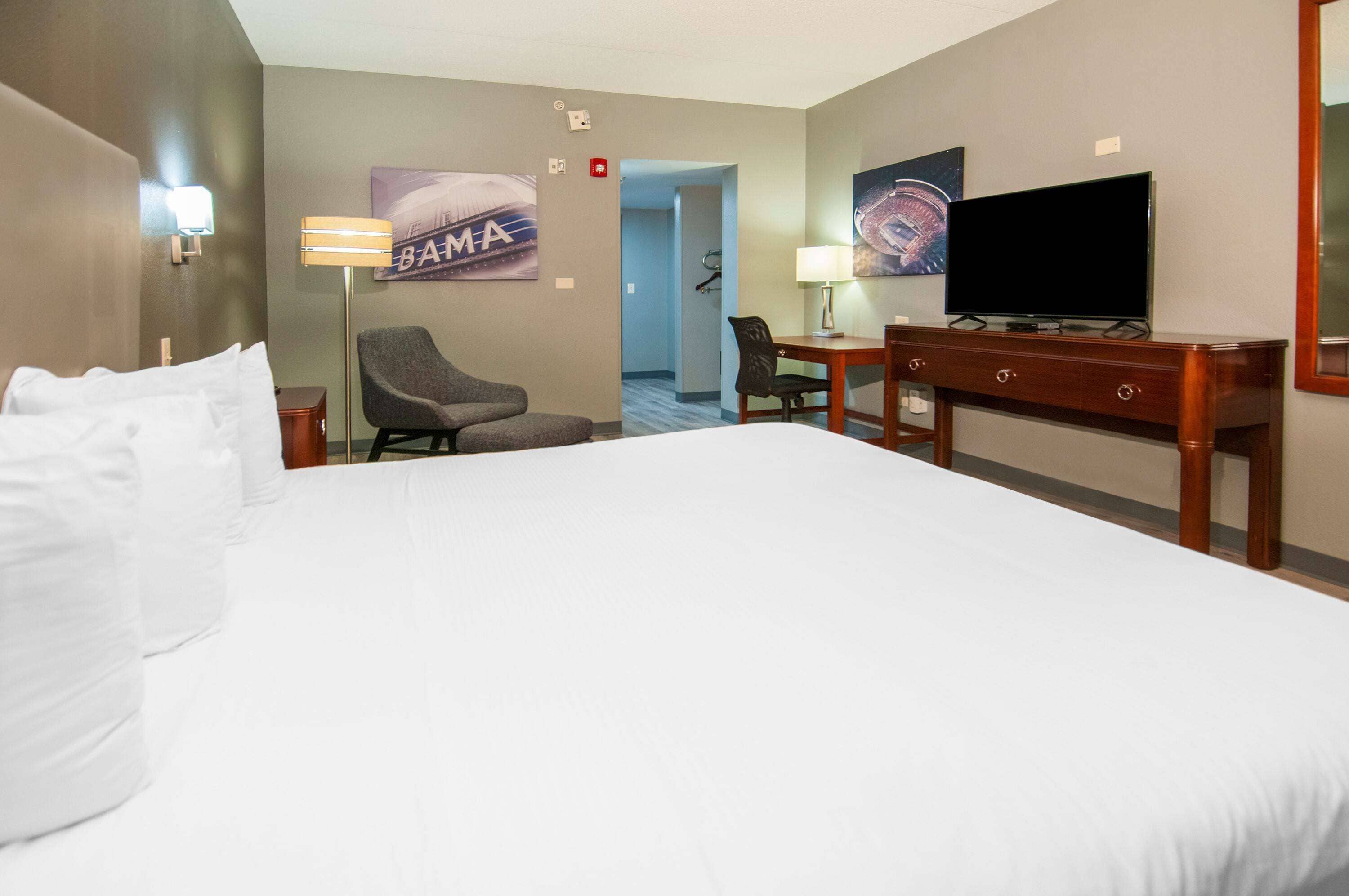 A hotel room with a large bed, a desk with a television, a chair, and a lamp.