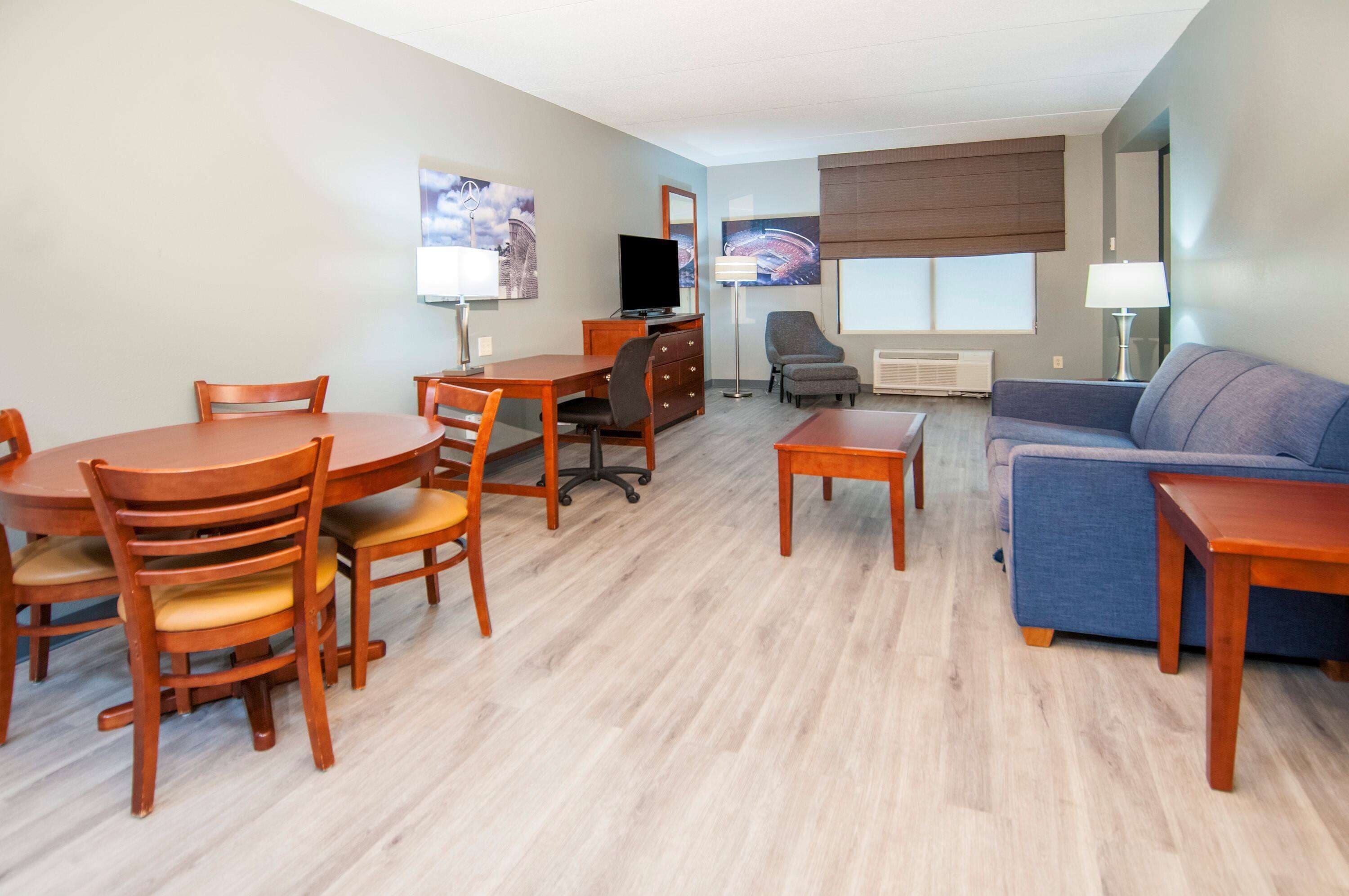 A hotel room with a dining area, a sofa, a television, and a large window with blinds.