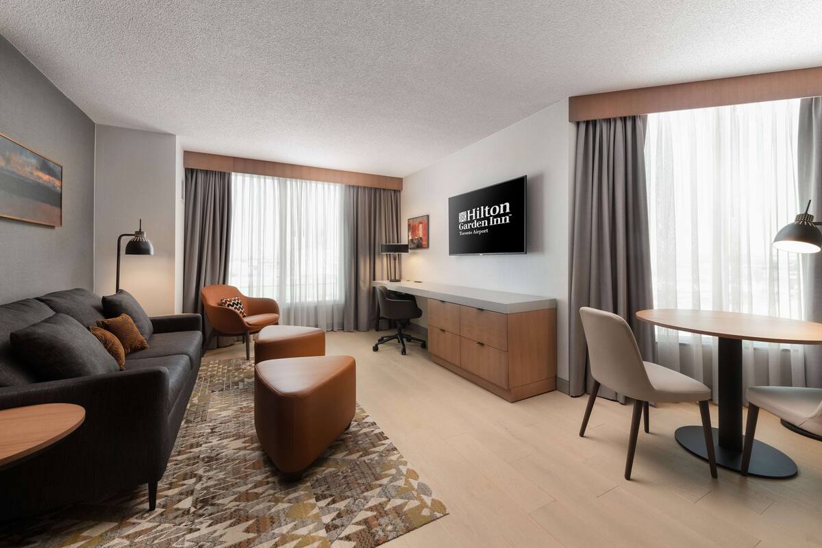 A modern hotel room with a sofa, ottoman, dining table, and a TV mounted on the wall.