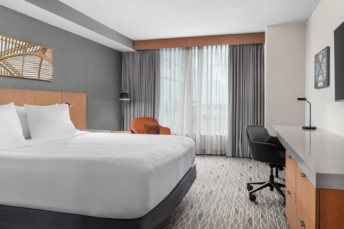 A hotel room with a large bed, a desk, an orange chair, and a view of the city through sheer curtains.