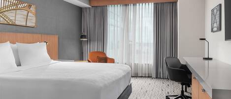A hotel room with a large bed, a desk, an orange chair, and a view of the city through sheer curtains.