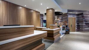 A modern reception area with wooden paneling and a counter, a refrigerator stocked with beverages, and a patterned wall in the background. - Hilton Garden Inn Toronto Airport (Mississauga)