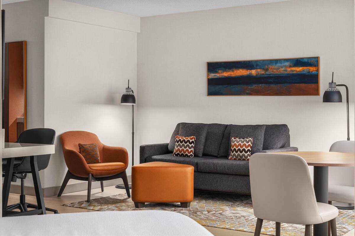 A modern living room with a grey sofa, orange armchair, and white chair. There is a painting on the wall, a black floor lamp, and a wooden table.