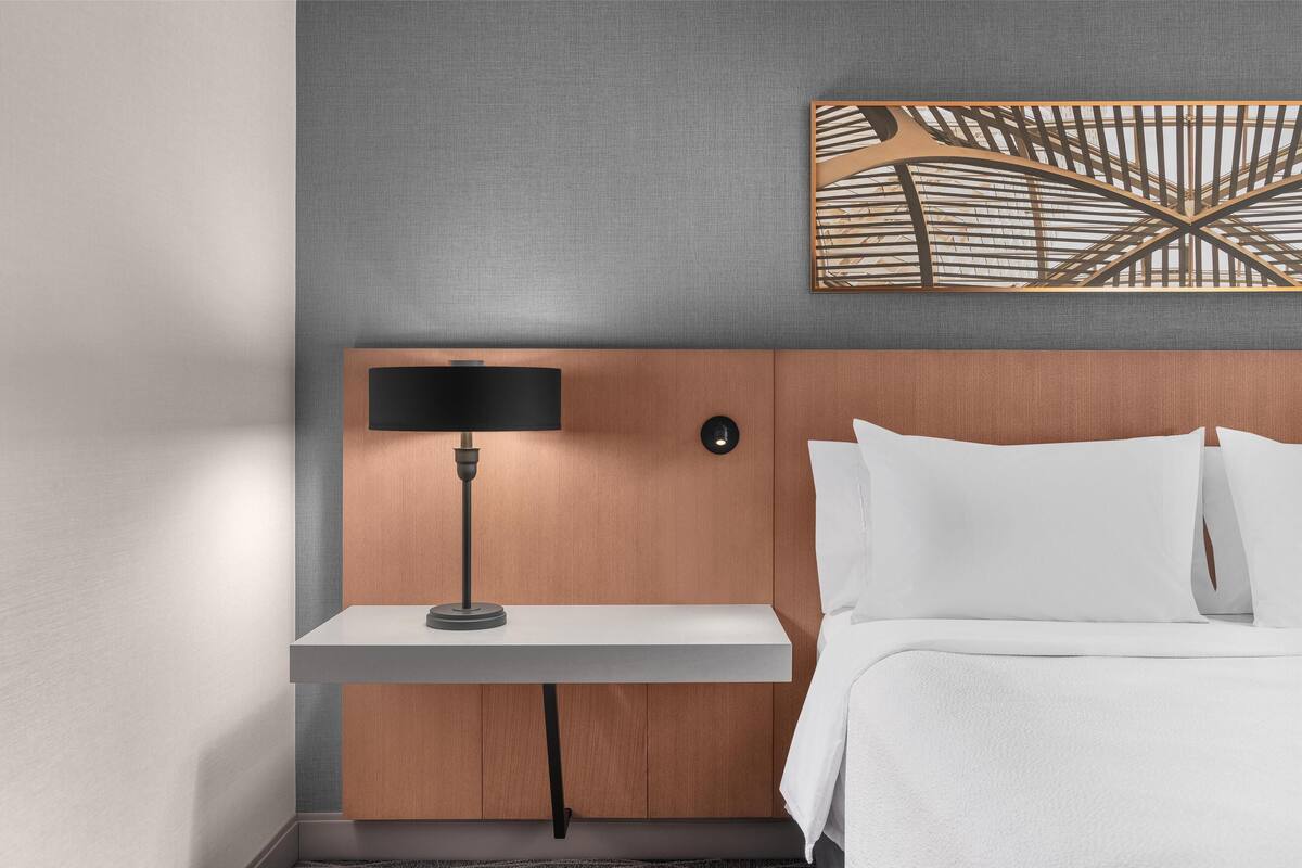 A hotel room with a wooden headboard, a bedside table with a lamp, and a painting above the bed.
