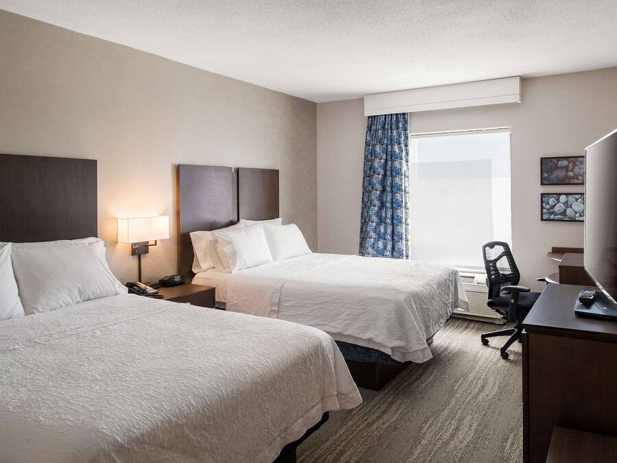 A hotel room with two beds, a desk with a TV, a chair, and a window with curtains.