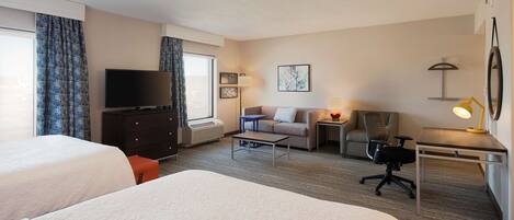 A hotel room with two beds, a TV, a desk with a lamp, a chair, a sofa, and a small table.