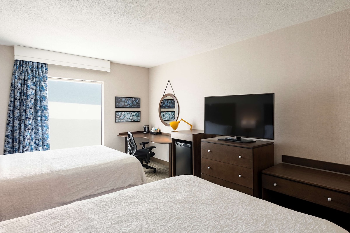 Ein Hotelzimmer mit zwei Betten, einem Schreibtisch mit Fernseher, einem Sessel und einer Schreibtischlampe.
