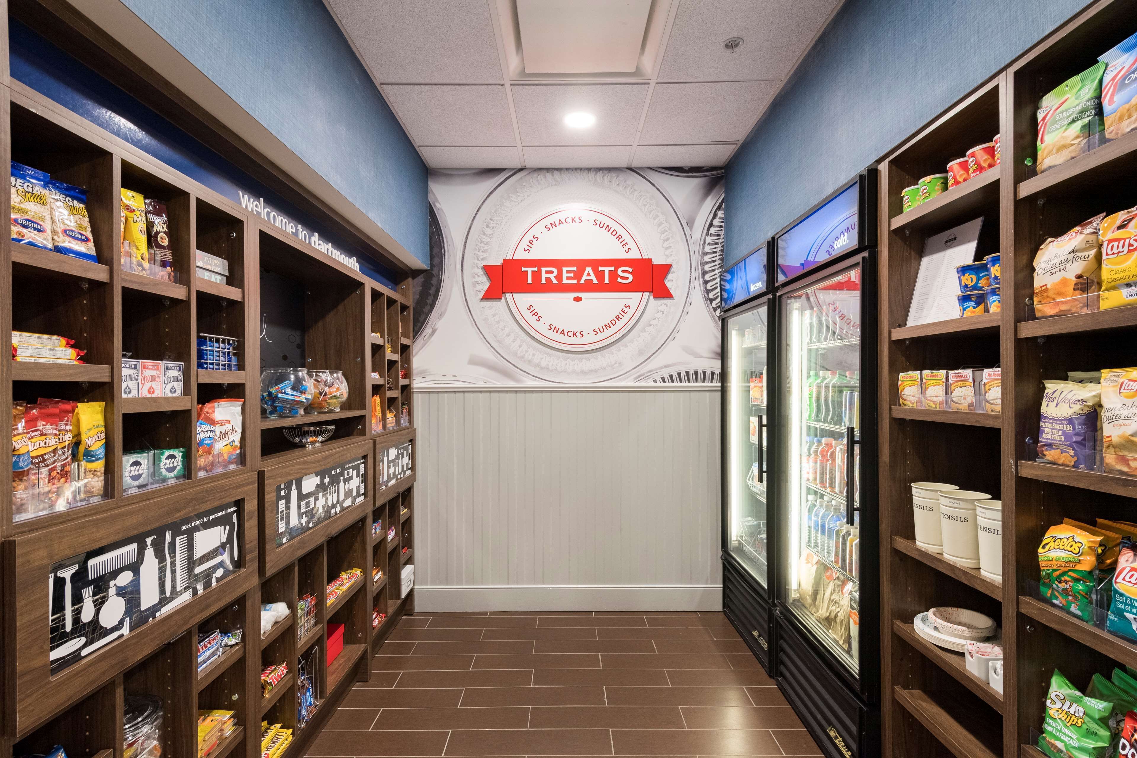 A corridor with shelves stocked with snacks and treats, a refrigerated display case, and a wall with a 'Treats' sign.