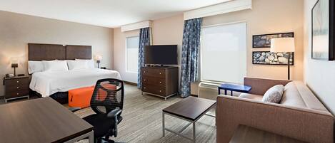 A hotel room with a bed, a desk, a chair, a sofa, a TV, a dresser, and artwork on the wall.