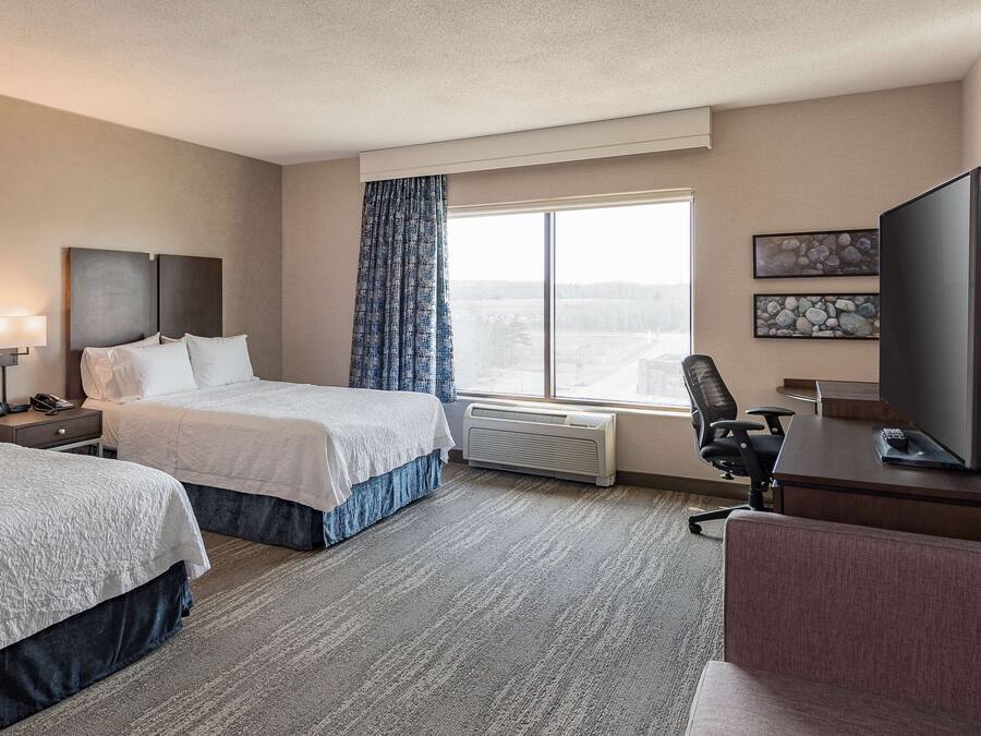 Ein Hotelzimmer mit zwei Betten, einem Schreibtisch, einem Sessel, einem Fernseher und einem großen Fenster mit Blick.
