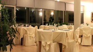 A banquet hall with round tables covered in white tablecloths, chairs with white covers, and a large window in the background. - Hotel Elvezia (Pesaro)