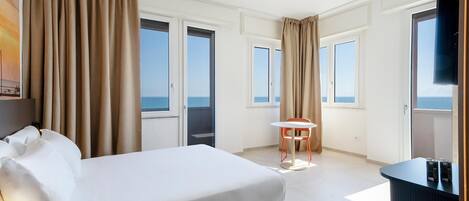A hotel room with a bed, a desk, a chair, and a view of the sea through large windows.