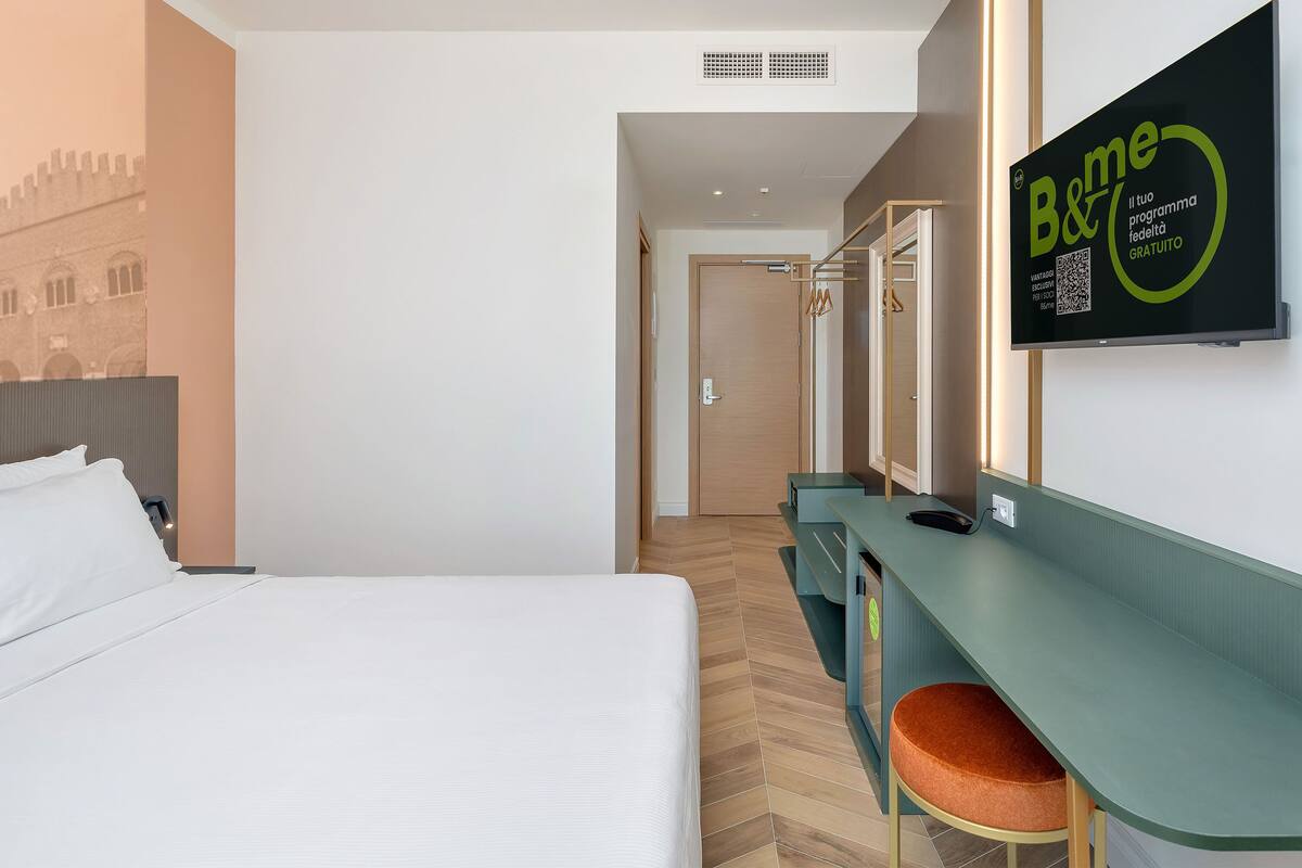 Une chambre d’hôtel comprenant un lit, un bureau avec une chaise, un miroir et un panneau fixé au mur.