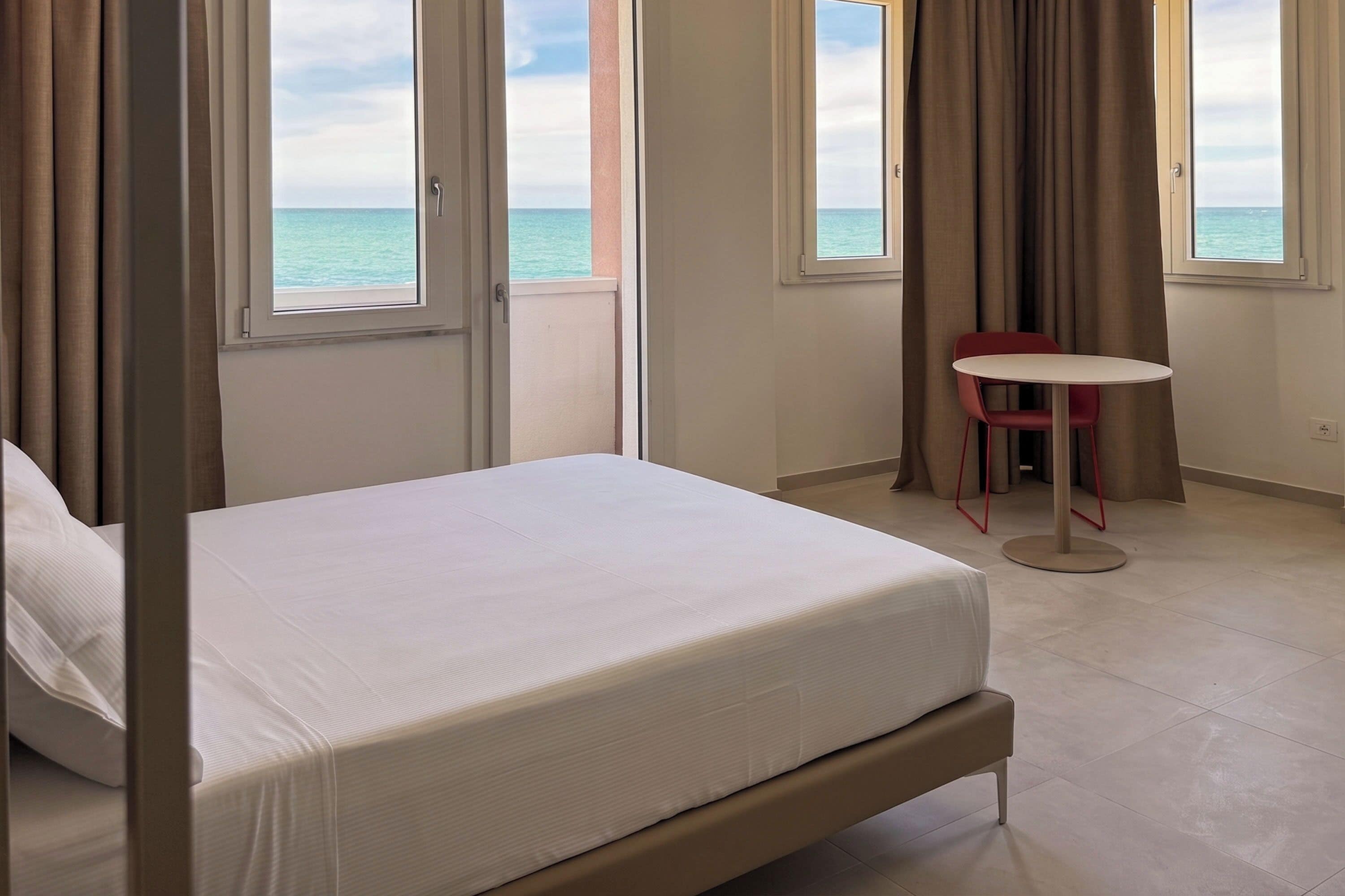 A hotel room with a bed, a small table, and a view of the sea through large windows.