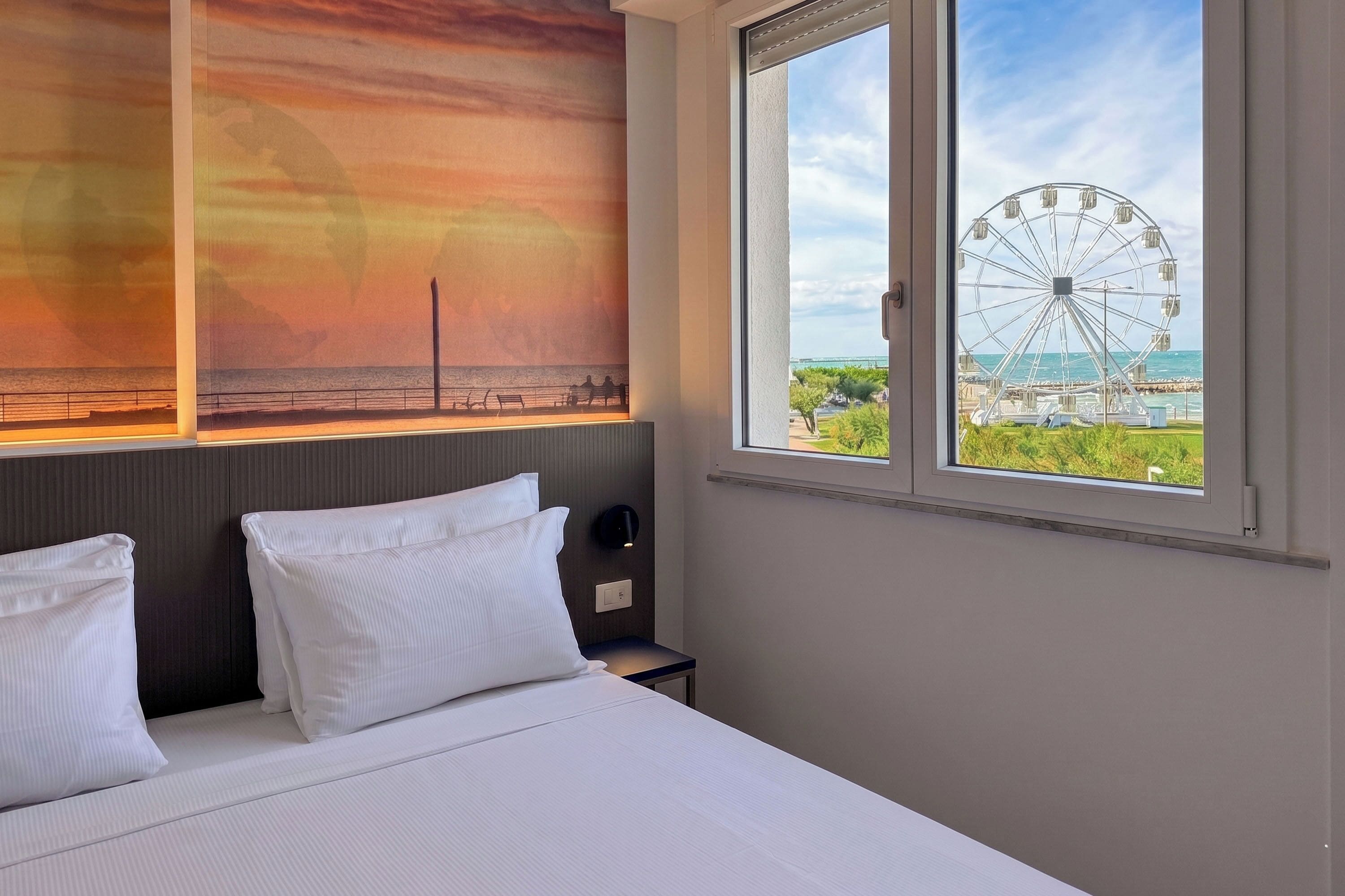 Ein Hotelzimmer mit einem Bett, Kissen, einem Nachttisch, einer Wandlampe und einem großen Fenster mit Blick auf einen Strand und ein Riesenrad.